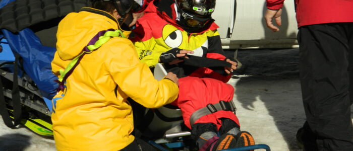 A Taos Ski Valley instructor kneels to assist a disabled skier in a ski chair in the parking lot. Two other skiers help.