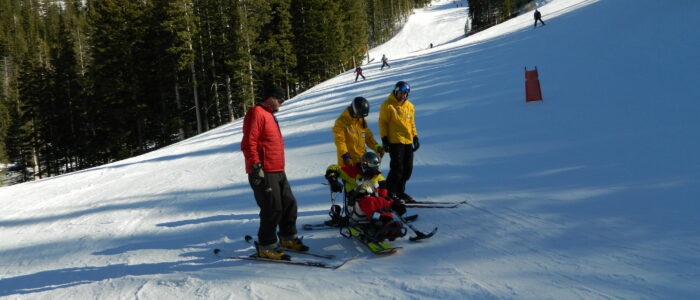 Two Taos Ski Valley instructors in yellow coats and a third skier assist a disabled skier in a ski chair on the slopes.