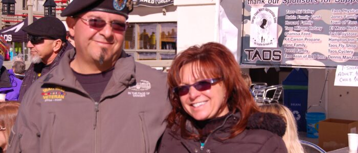 A veteran and companion embrace side by side at the foot of Taos Ski Valley. Veteran-appreciation and sponsor banners behind.