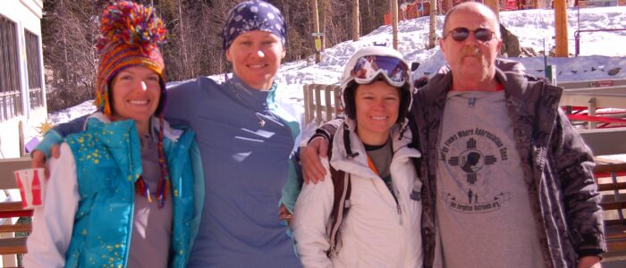 Four cheerful skiers embrace side by side under the sun. Patio and wintry forest in the background. Lodge in the distance.