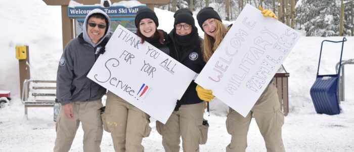 Four young adults in cargo pants and black boots at Taos Ski Valley holding signs welcoming and thanking military families.