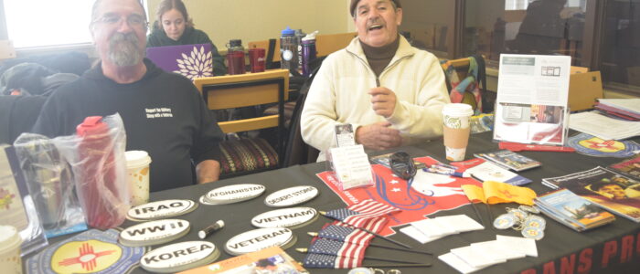 Two cheerful men sit behind a table with veteran brochures, patches, bumper stickers, keychains, and flags