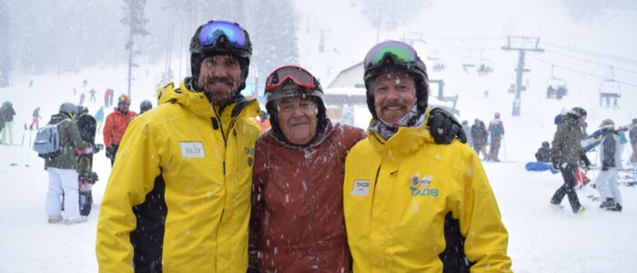 A older skier embraces two Taos Ski Valley instructors. Chairlift, skiers, and snowboarders in background. Snowing heavily.