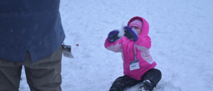 Child skier in pink coat and hat plays in the snow, other skiers standing around