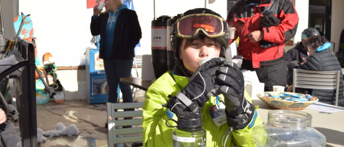 Young skier in bright yellow jacket and gloves sipping a drink at a donation table at Taos Ski Valley, other skiers milling