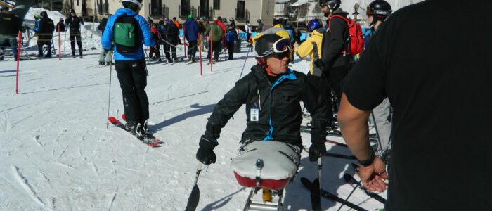 Disabled skier in ski chair at Taos Ski Valley amidst crowd of skiers milling around entrance to chairlift. Lodges behind.