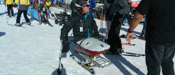 Disabled skier in ski chair at Taos Ski Valley base amidst many other skiers. Rhonda's restaurant complex in background.