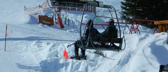A disabled skier in a ski chair and companion ride a chairlift near the base of Taos Ski Valley on a sunny day.