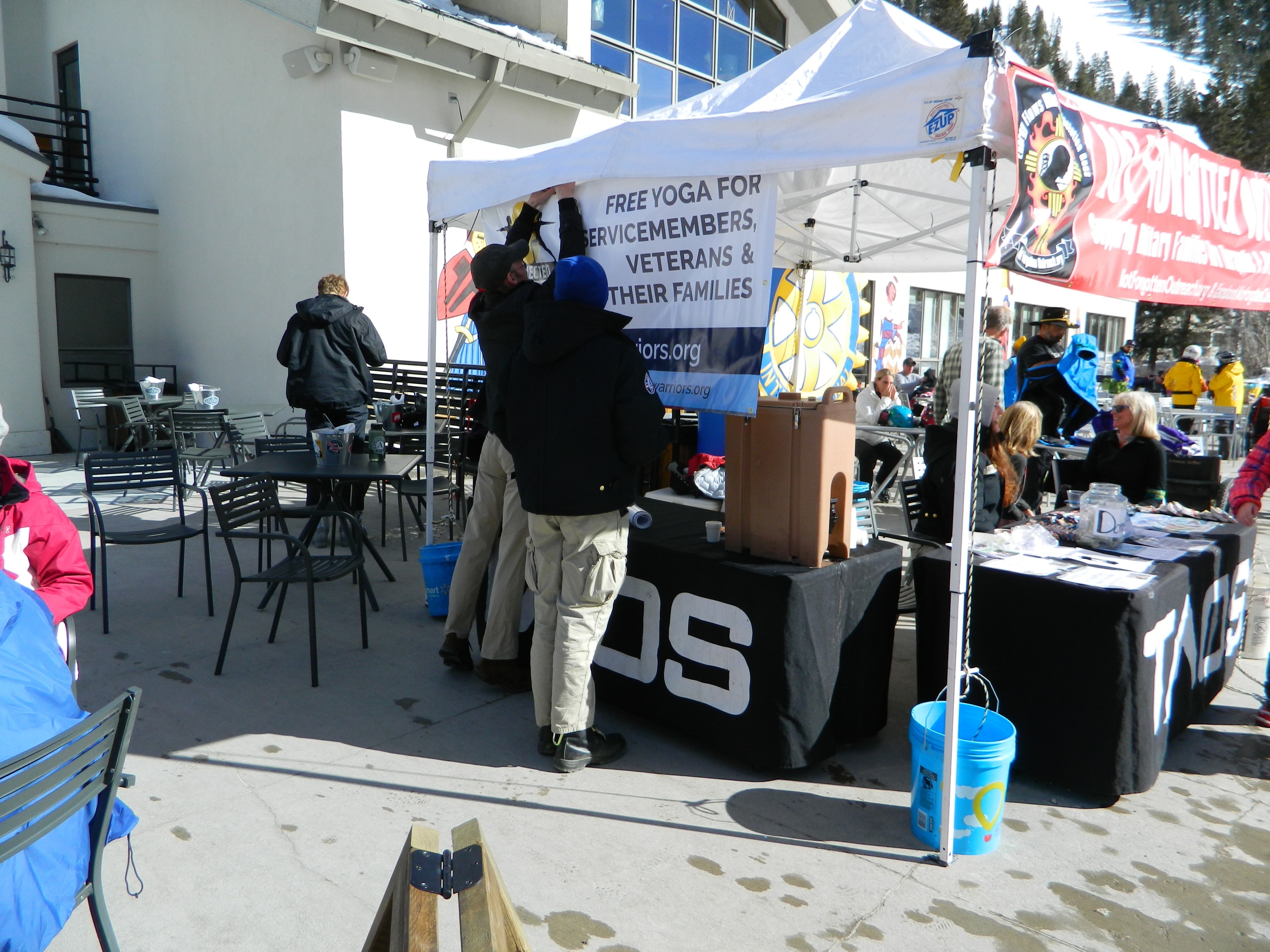 Staff fixes yoga banner on white Not Forgotten Outreach tent at the base of Taos Ski Valley in front of lodge.