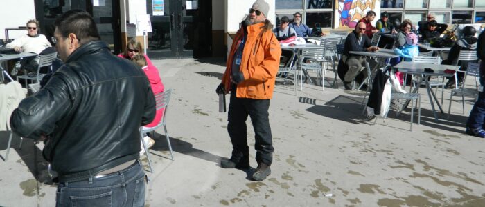 A man in an orange jacket standing on the patio in front of Tenderfoot Katie's at Taos Ski Valley. Others relaxing behind.