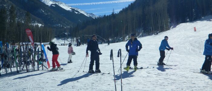 A pair of skis and poles by themselves at the base of Taos Ski Valley. Skiers, wintry forest, mountain peaks, blue skies.