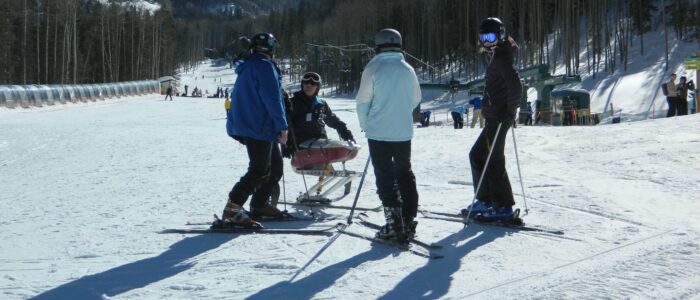 Four skiers, one disabled, confer at the base of Taos Ski Valley. Peaks, wintry forest, skiers, and lodge behind. Blue skies.