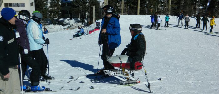 Group of skiers, one disabled, confer on the slopes of Taos Ski Valley. Other skiers behind, and main lodge in the distance.