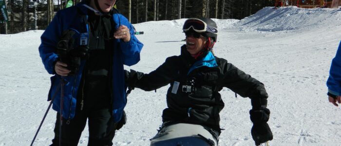 Laughing disabled skier in ski chair jokes with another skier on the slopes at Taos Ski Valley. Wintry forest behind.