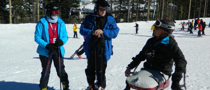 Three skiers, one of them disabled, confer solemnly on the slopes at Taos Ski Valley. Skiers, forest, and lodge behind.