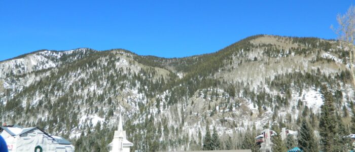 Bright blue sunny day at Taos Ski Valley village, seen from afar. Big crowd of skiers milling near Tenderfoot Katie's patio.