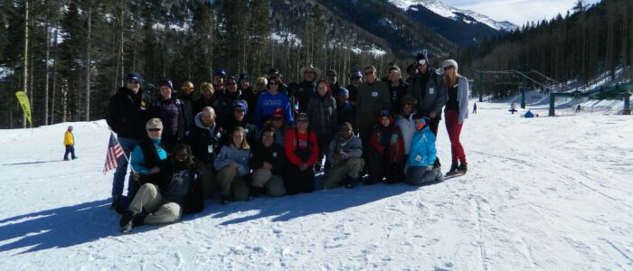 Not Forgotten Outreach group photo at the base of Taos Ski Valley. Chairlift, mountain peaks, blue sky, wintry forest behind.