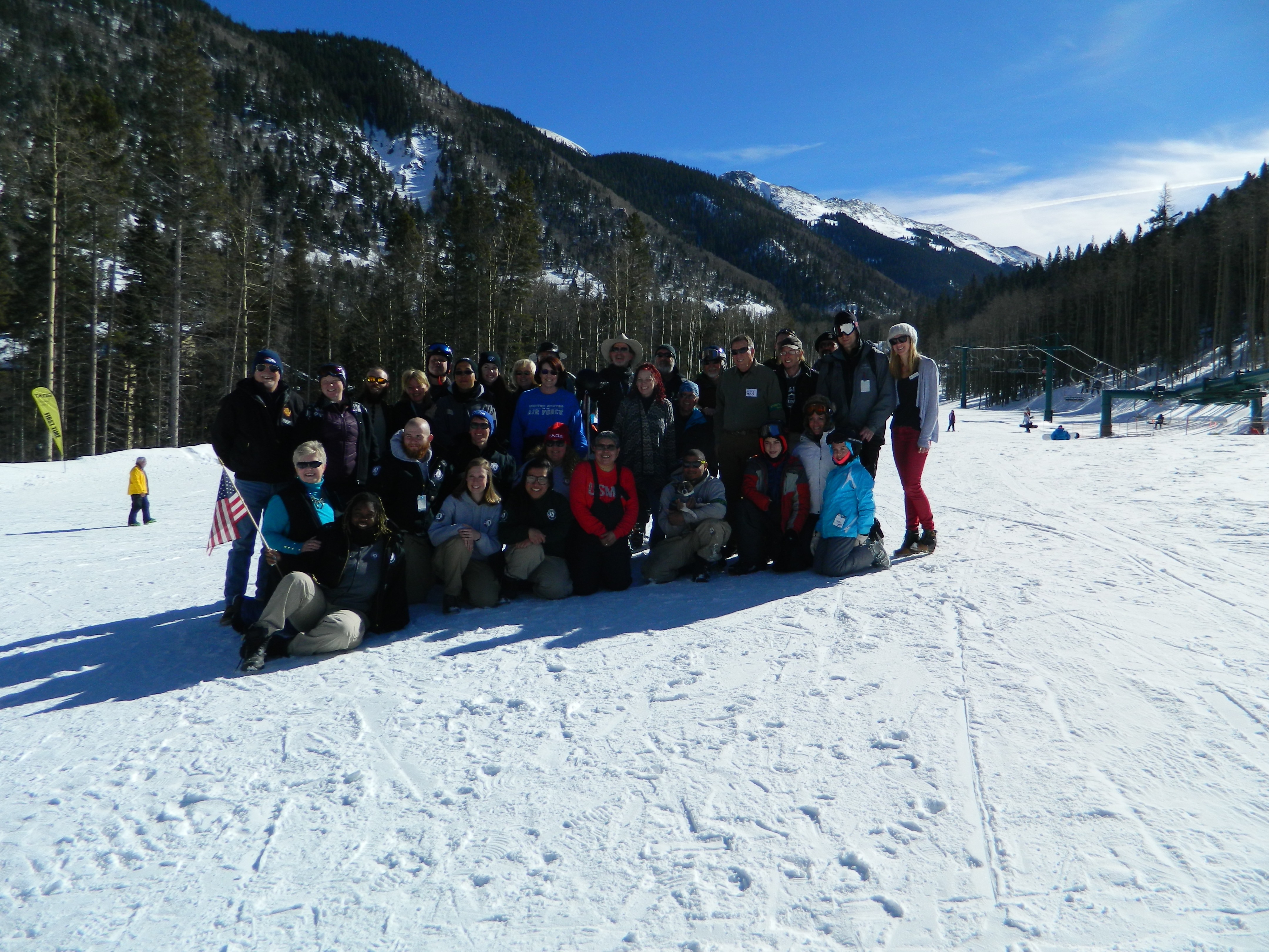Not Forgotten Outreach group photo at the base of Taos Ski Valley. Chairlift, mountain peaks, blue sky, wintry forest behind.