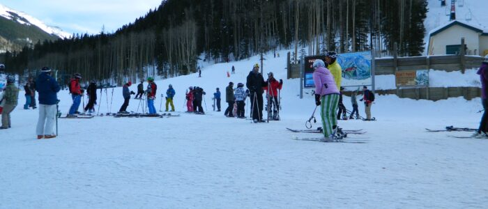 Many skiers conversing at the foot of Taos Ski Valley in the late afternoon shade.
