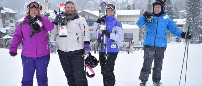 Four cheerful skiers at Taos Ski Valley on a snowy day holding their ski equipment over their shoulders. Lodge behind.