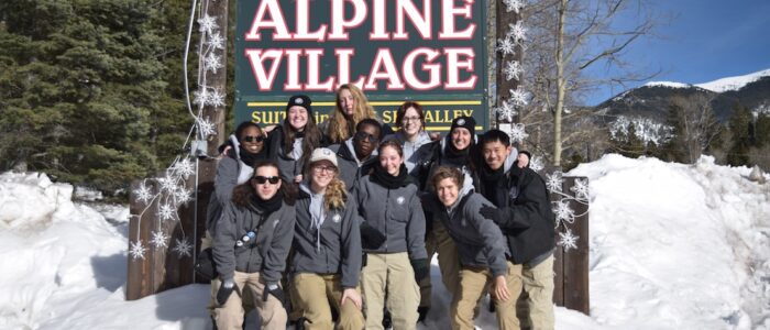 Eleven young adults in two rows posing in the snow bank in front of the Alpine Village sign at Taos Ski Valley, blue skies