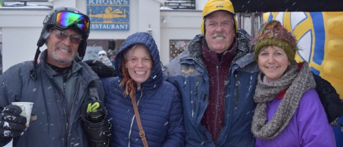 One skier with boots over shoulder joining three others in side-by-side embrace near Rhoda’s Restaurant in Taos Ski Valley